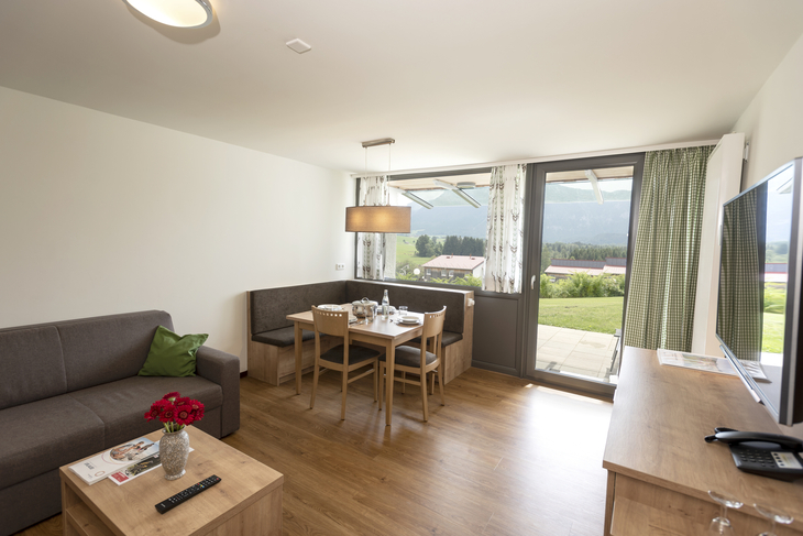 Wohn-Essbereich in der Ferienanlage Inzell, mit Holzfußboden, Esstisch mit Eckbank und Stühlen, Sofa mit kleinem Tisch, Sideboard mit Fernseher darüber und großem Fenster mit Tür zur Terrasse.