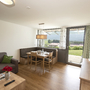 Wohn-Essbereich in der Ferienanlage Inzell, mit Holzfußboden, Esstisch mit Eckbank und Stühlen, Sofa mit kleinem Tisch, Sideboard mit Fernseher darüber und großem Fenster mit Tür zur Terrasse.