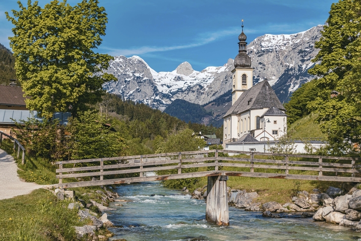 Kirche St. Sebastian in Ramsau vor Alpenkulisse und Holzbrücke über Fluss.
