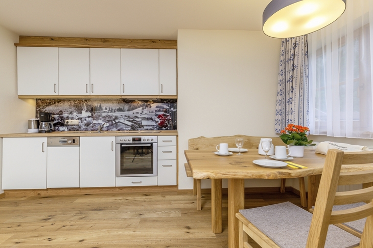 Küche in der Ferienanlage Oberwössen, mit Holzfußboden, heller Küchenzeile und gedecktem rustikalen Holzesstisch mit Bank und zwei Stühlen vor einem Fenster mit blau-weißem Vorhang.