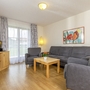 Wohnzimmer in der Ferienanlage Oberwössen, mit Holzfußboden, grauem Sofa und zwei Sesseln, einem kleinen Holztisch und einem Fernseher auf einem Sideboard, sowie einem bodentiefen Fenster mit blau-weißen Vorhängen.