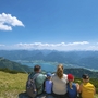Familie sitzt auf Wiese mit Blick auf See und Berge unter blauem Himmel.