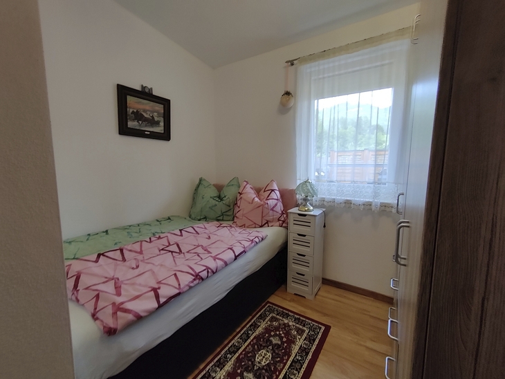 Kleines Schlafzimmer mit Einzelbett, Fenster mit Vorhang, Wandbild und Teppich.
