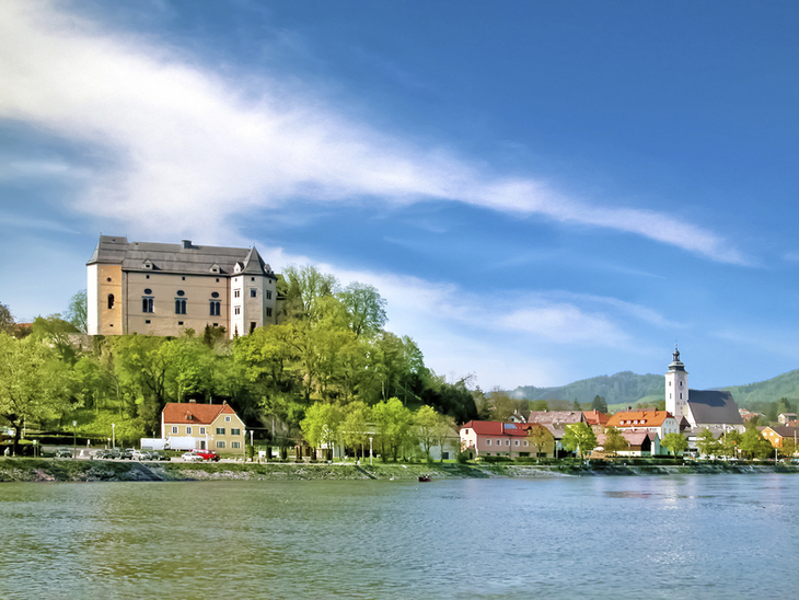 Blick auf eine Burg und eine Kirche am Ufer der Donau bei blauem Himmel.