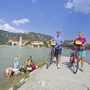 Radfahrer auf einem Steg am Donau-Radweg in der Wachau, im Hintergrund Berge und Kirche.