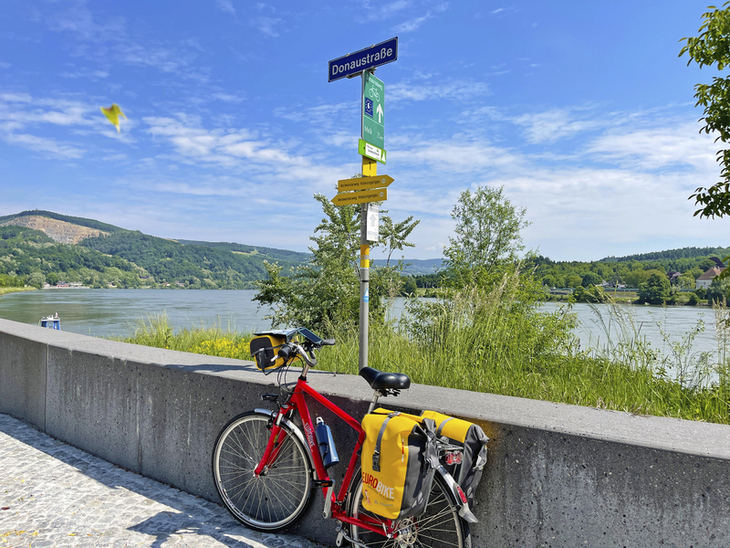 Rotes Fahrrad vor Donau und Radwegweiser unter blauem Himmel.