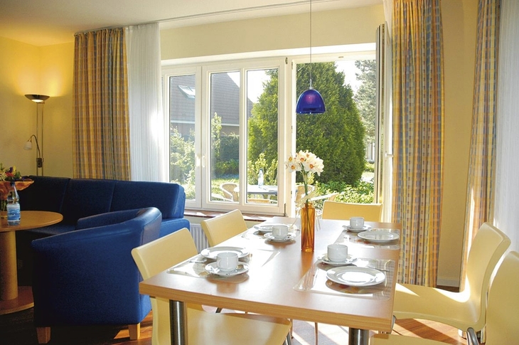 Wohnbereich mit Esstisch, blauen Sesseln und Blick auf Garten in Harlesiel-Ferienanlage.