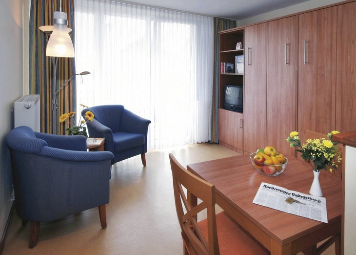 Wohnzimmer einer Ferienwohnung mit Sessel, Tisch, Fernseher und Blumendekoration.