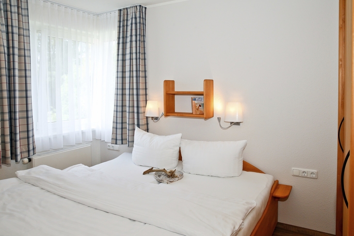 Schlafzimmer mit Doppelbett, Fenster mit Vorhang und Wandregal mit Dekoration.