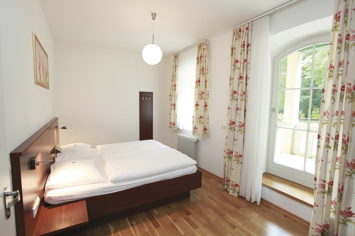 Helles Schlafzimmer mit Doppelbett, Holzboden und Fenster mit Vorhängen.