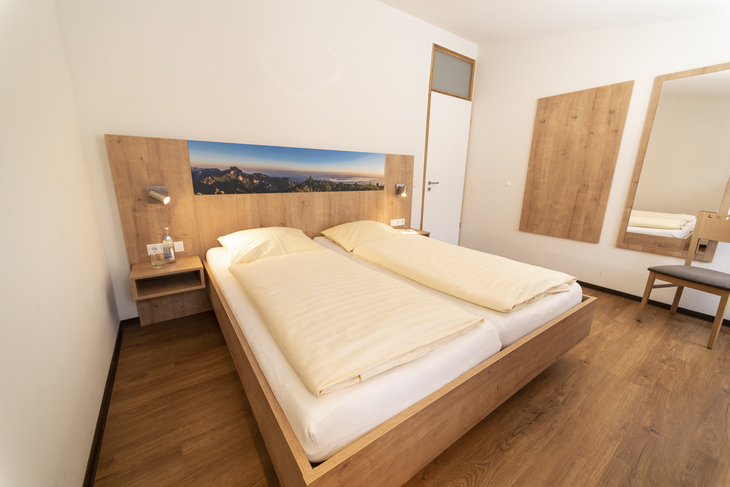 Schlafzimmer in der Ferienanlage Inzell, mit Holzfußboden, Doppelbett mit Nachttischen, Lampen und Bergpanoramabild am Kopfteil.