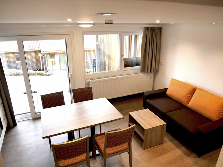 Wohnzimmer mit Esstisch, Stühlen, Sofa und großen Fenstern mit Blick nach draußen.