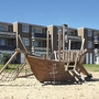 Spielplatz in Form eines Piratenschiffs aus Holz, vor den dreistöckigen Reihenhäusern mit Balkonen, der Ferienanlage Cadzand-Bad unter strahlend blauem Himmel.