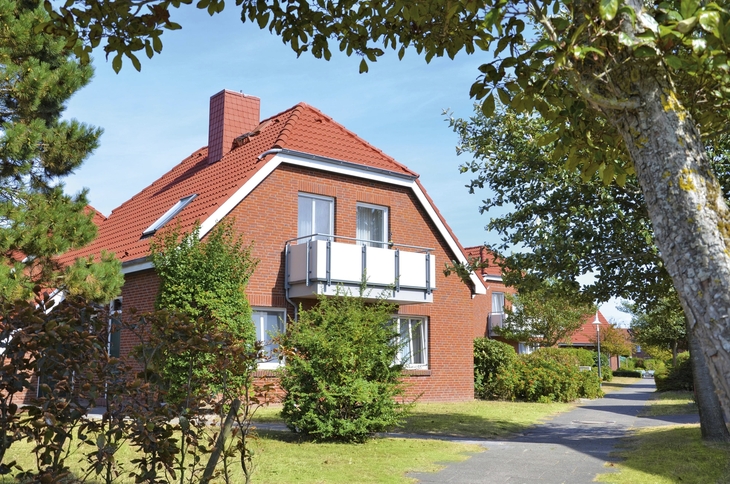 Rotes Ferienhaus mit Garten und Balkon in Ferienanlage Harlesiel, umgeben von grünen Bäumen und einem Gehweg.
