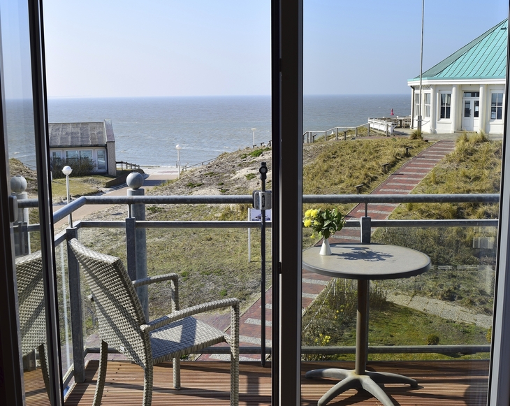 Blick vom Balkon, mit Tisch und Stuhl, der Ferienanlage Norderney auf das Meer. Rechts im Bild der achteckige weiße Pavillon mit grünem Dach.