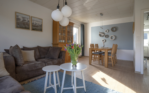Wohnzimmer mit Sofa, zwei Tischen, Blumenstrauß, Esstisch und Stühlen in hellem Raum.