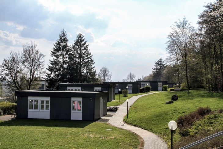 Pfad führt zu modernen Ferienhäusern mit Flachdach, weißen Türen und Fenstern sowie anthrazitfarbener Fassade, der Ferienanlage Prüm in grüner, bewaldeter Landschaft. unter leicht bewölktem Himmel