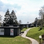 Pfad führt zu modernen Ferienhäusern mit Flachdach, weißen Türen und Fenstern sowie anthrazitfarbener Fassade, der Ferienanlage Prüm in grüner, bewaldeter Landschaft. unter leicht bewölktem Himmel