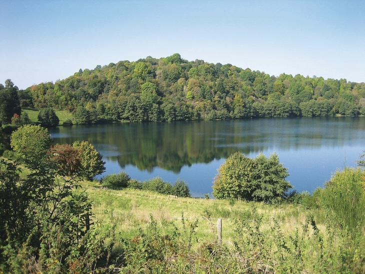 Blick auf einen See umgeben von Wiesen und Büschen mit bewaldetem Hügel im Hintergrund unter blauem Himmel mit Sonnenschein.