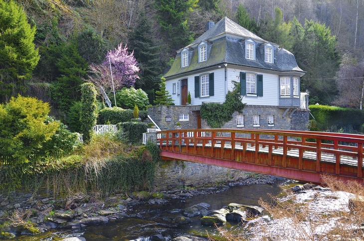 Ein Haus in Monschau mit Brücke über einen Bach und umgeben von Bäumen.