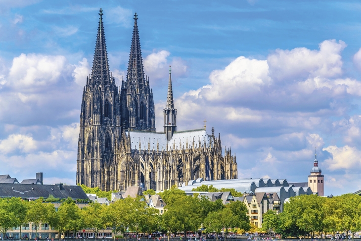 Kölner Dom vor blauem Himmel mit Wolken und grünen Bäumen im Vordergrund.