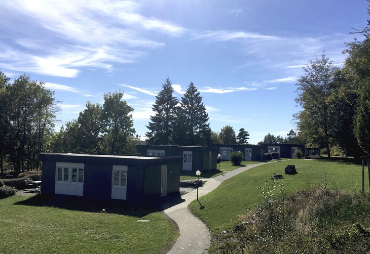 Mehrere kleine Gebäude in einer grünen Parklandschaft bei sonnigem Wetter.
