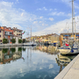 Segelboot im Hafen von Grado vor bunten Gebäuden und klarem Himmel.
