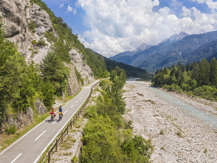 Radfahrer auf einem Weg entlang eines Flusses in einer bergigen Landschaft.
