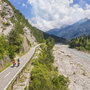 Radfahrer auf einem Weg entlang eines Flusses in einer bergigen Landschaft.