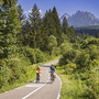 Zwei Radfahrer auf einem Waldweg mit Bergpanorama im Hintergrund.
