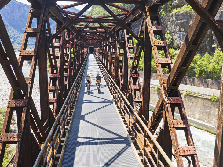 Radfahrer auf einer Metallbrücke über einem Fluss in einer bergigen Landschaft.
