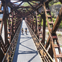 Radfahrer auf einer Metallbrücke über einem Fluss in einer bergigen Landschaft.