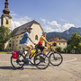 Zwei Radfahrer fahren auf einem Platz, im Hintergrund eine Kirche und Berge.