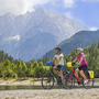 Zwei Personen fahren Fahrrad am See mit Bergen im Hintergrund.