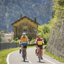 Zwei Radfahrer auf einem Radweg vor einem Haus bei sonnigem Wetter.