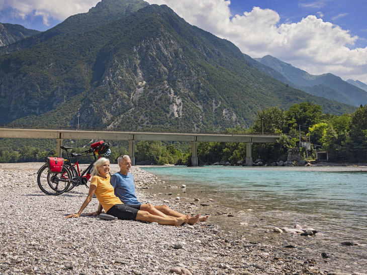 Zwei Personen sitzen an einem Flussufer mit Fahrrädern und Blick auf Berglandschaft.