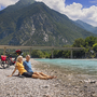 Zwei Personen sitzen an einem Flussufer mit Fahrrädern und Blick auf Berglandschaft.