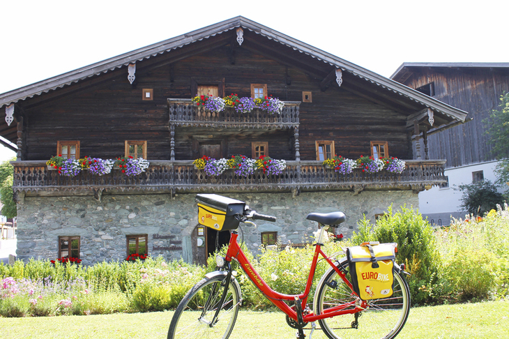 Rotes Fahrrad vor traditionellem Chalet mit blumengeschmücktem Balkon im Sommer.