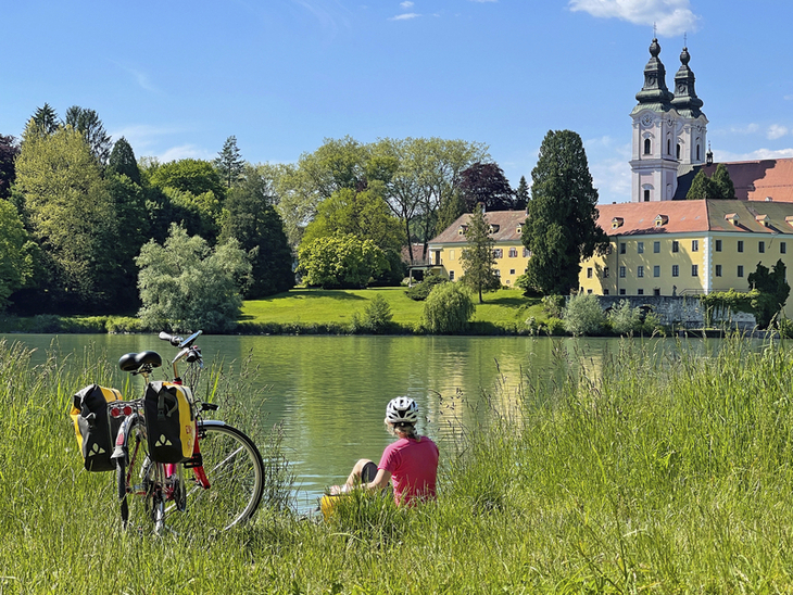 Radfahrer macht Pause am Flussufer mit Blick auf eine Kirche und Gebäude im Hintergrund.