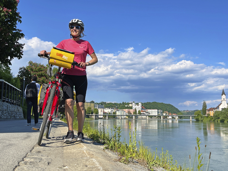Fahrradfahrerin am Ufer eines Flusses mit Landschaft im Hintergrund auf dem Tauernradweg.