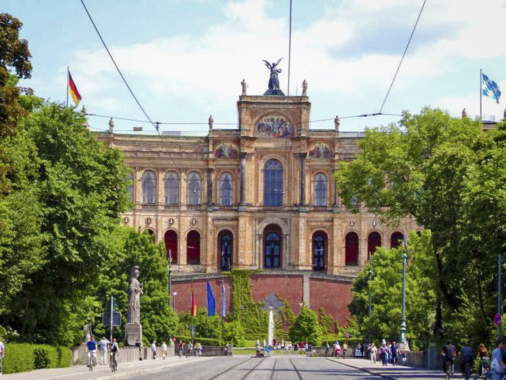 Münchner Maximilianeum mit Flaggen, Bäume und Straße im Vordergrund bei Sonnenschein.