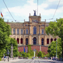Münchner Maximilianeum mit Flaggen, Bäume und Straße im Vordergrund bei Sonnenschein.