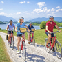Gruppe von Radfahrern auf einem Weg vor einem See und den Alpen im Hintergrund.