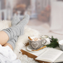 Füße in grauen Socken auf Fell, Tee und Buch im Hintergrund, winterlich gemütlich.