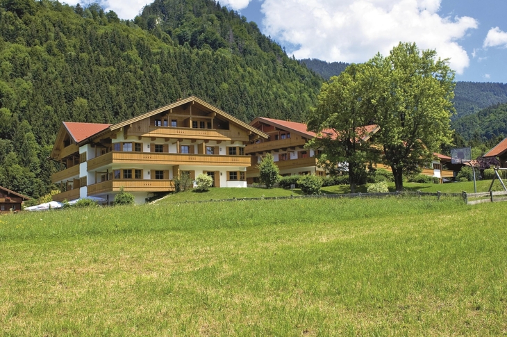 Holzgebäude vor bewaldeten Hügeln mit grünem Feld im Vordergrund.