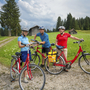Drei Radfahrer in einer ländlichen Landschaft mit Wiesen und Hütten.