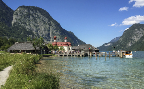 See mit Kirche St. Bartholomä und Bergen im Hintergrund bei sonnigem Wetter.