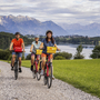 Drei Radfahrer auf einem Schotterweg am See mit Bergpanorama im Hintergrund.