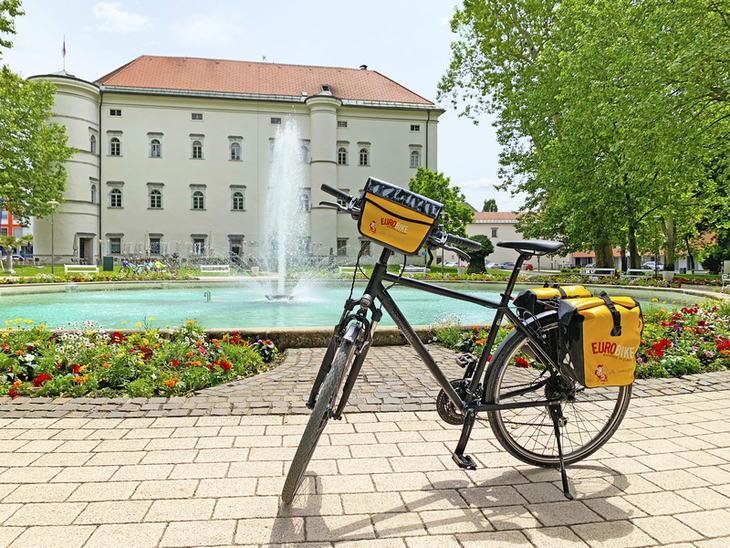 Fahrrad vor historischem Gebäude und Brunnen mit Blumenbeet im Vordergrund.