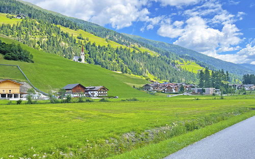 Landschaft mit Radweg, grünen Wiesen, Dorf und Kirche in den Alpen
